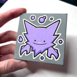 A hand holding a mini canvas featuring Ditto Haunter with white outlines and small purple bubbles, photographed against a light background.