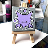 A mini canvas painting of a Ditto Haunter with a white outline, displayed on a mini wooden easel. The background is a blurred art desk.