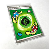 A Pokémon TCG Basic Energy card featuring a green background with a central leaf icon and cartoon creatures, sealed in a clear protective sleeve and photographed on a white surface.