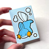 A hand holding a small rectangular canvas painting featuring a Ditto Mudkip with white outlines, set against a plain light background.