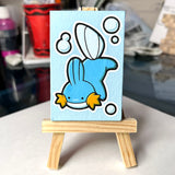 A small canvas painting of a Ditto Mudkip, displayed on a miniature wooden easel against a blurred indoor background.