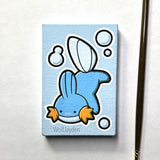 A small rectangular canvas painting featuring a Ditto Mudkip with white outlines, photographed on a white background. A paintbrush is visible along the right edge.